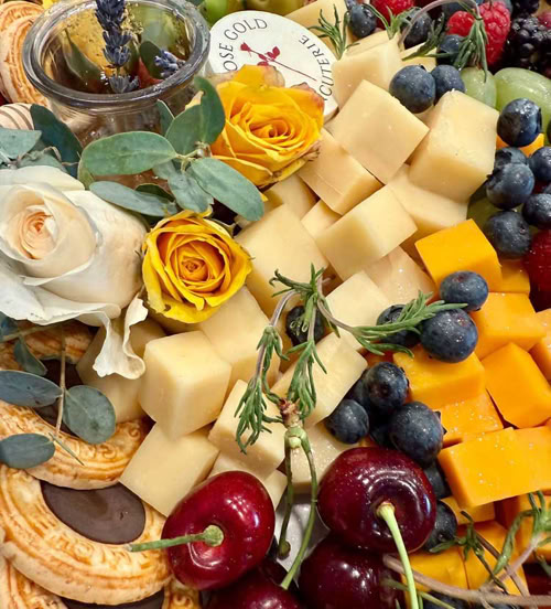 Colorful charcuterie board with cheese cubes, berries, cherries, cookies, flowers, and a honey jar, branded by Rose Gold Charcuterie.