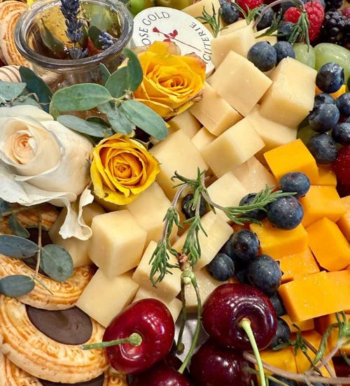 Colorful charcuterie board with cheese cubes, berries, cherries, cookies, flowers, and a honey jar, branded by Rose Gold Charcuterie.