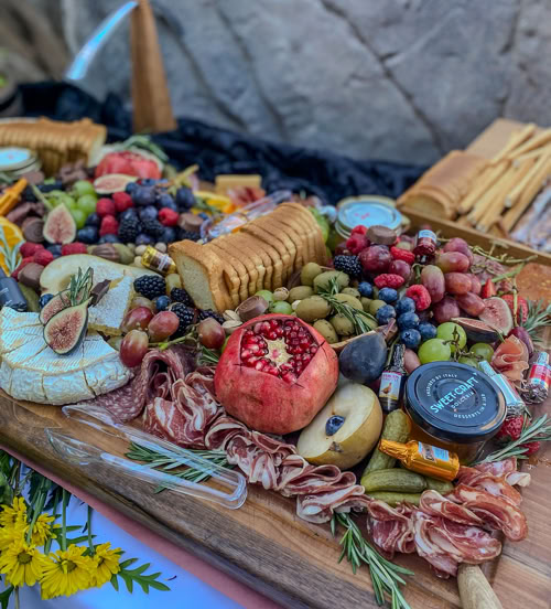 Event grazing table with gourmet cheeses, pomegranate, pickles, crackers, meats, and olives styled with rosemary and fresh fruit by Rose Gold Charcuterie.