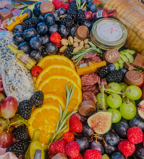 A colorful charcuterie board filled with an assortment of fresh fruits (blackberries, grapes, raspberries, figs, orange slices), cured meats, cheese log with poppy seeds, chocolate candies, mini honey jars, nuts, rosemary sprigs, and a tin labeled “Rose Gold Charcuterie.”