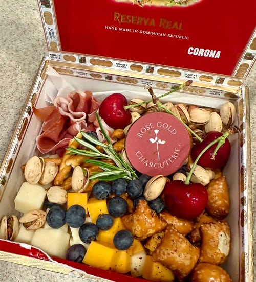 Unique cigar box charcuterie by Rose Gold Charcuterie featuring cubes of cheese, blueberries, cherries, pistachios, pretzels, cured meat, and a branded label inside a Romeo y Julieta cigar box.