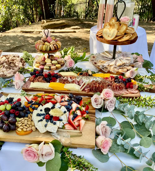 A vibrant charcuterie spread featuring two large round trays and a rectangular wooden board, all filled with an assortment of colorful fresh fruits like strawberries, grapes, and blueberries, assorted cheeses, meats, crackers, chocolates, and chocolate-covered pretzels. The display includes garnishes of rosemary and figs, creating a rich and inviting presentation suitable for a party or upscale event.  