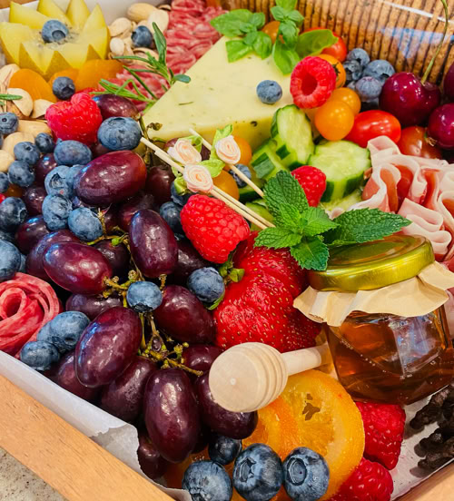 Colorful charcuterie grazing box filled with grapes, berries, cheese, cured meats, fresh herbs, and honey dipper.