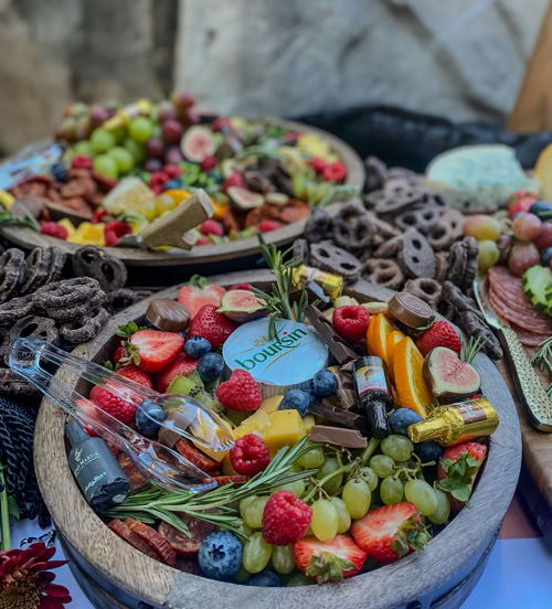 A vibrant round charcuterie board filled with assorted fresh fruits (grapes, blueberries, raspberries, figs, orange slices), Boursin cheese, chocolate pieces, chocolate-covered pretzels, salami, rosemary sprigs, and mini honey jars. Other overflowing charcuterie trays are visible in the background.