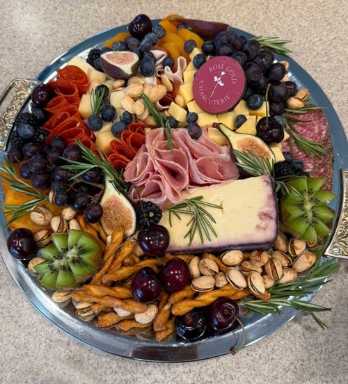 Round charcuterie platter with meats, cheeses, figs, cherries, kiwi, pretzels, pistachios, and a Rose Gold Charcuterie label in the center.