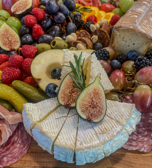 Rose Gold Charcuterie spread with creamy brie cheese topped with fresh figs and rosemary, surrounded by grapes, berries, pickles, artisan bread, and assorted meats—perfect for upscale grazing tables and event catering.