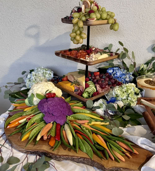 A beautifully arranged table featuring an assortment of food, with a floral centerpiece and various cut flowers adorning the setting. The indoor scene highlights a decorative wall in the background, enhancing the still life presentation from Rose Gold Charcuterie in the Inland Empire