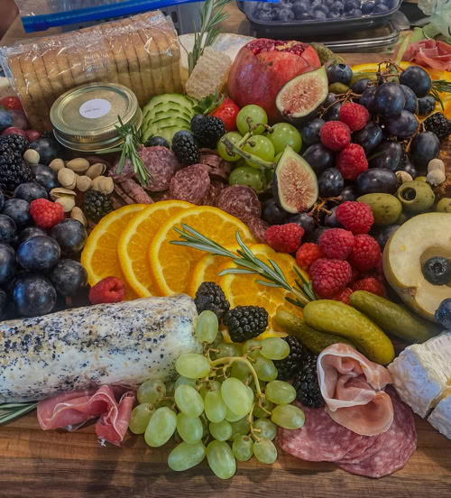 Colorful Rose Gold Charcuterie board loaded with gourmet cheeses, green grapes, fresh figs, pomegranate, orange slices, crackers, honeycomb, and artisan meats—styled for luxury grazing and special event catering.