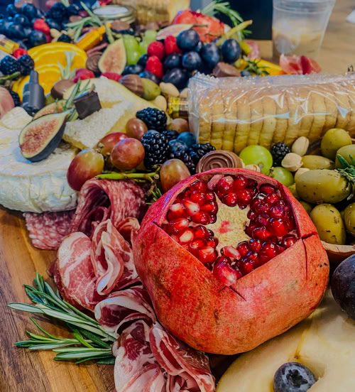 Artisan charcuterie board by Rose Gold Charcuterie featuring a halved pomegranate, brie, salami roses, fresh figs, grapes, berries, olives, and crackers—perfect for luxury grazing tables and holiday entertaining.
