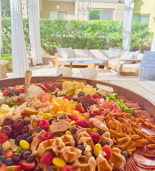 Large grazing table filled with cheeses, cured meats, waffles, pretzels, fresh berries, grapes, cucumbers, and sweet treats on a covered patio.