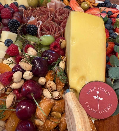 Charcuterie board with a large cheese wedge, salami roses, cherries, grapes, berries, pistachios, pretzels, and a Rose Gold Charcuterie label.