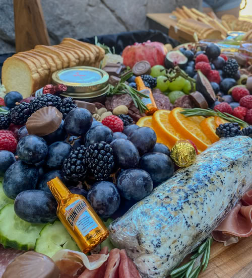 A gourmet charcuterie board featuring black grapes, blackberries, orange slices, cucumber, prosciutto, chocolate candies, mini liquor bottles, a herb-crusted cheese log, crackers, green grapes, and a branded tin from Rose Gold Charcuterie. The display is artfully arranged with pops of color and fresh rosemary.          A gourmet charcuterie board featuring black grapes, blackberries, orange slices, cucumber, prosciutto, chocolate candies, mini liquor bottles, a herb-crusted cheese log, crackers, green grapes, and a branded tin from Rose Gold Charcuterie. The display is artfully arranged with pops of color and fresh rosemary. 