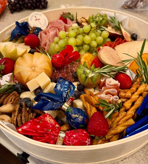 Round charcuterie box with meats, cheeses, grapes, pears, kiwi, berries, crackers, wrapped candies, and a Rose Gold Charcuterie label.