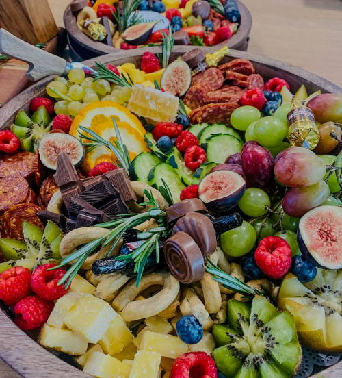 Round grazing platter featuring fresh figs, kiwi, berries, chocolate, citrus, cucumbers, and charcuterie meats—perfect for events and parties.