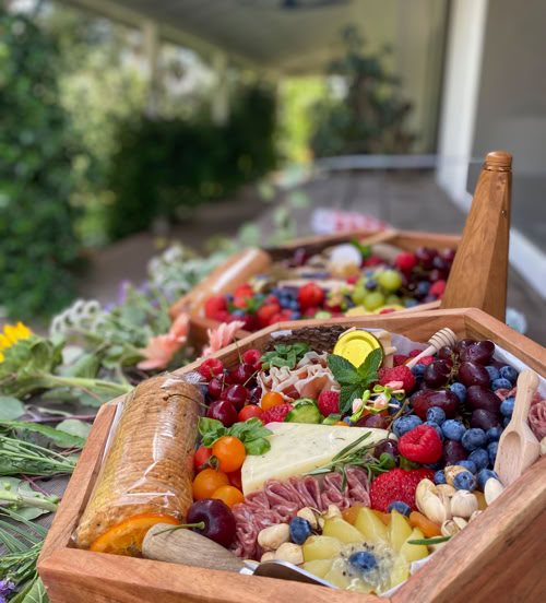 A wooden charcuterie box overflowing with fresh fruits (grapes, blueberries, strawberries, tomatoes), cheeses, meats, crackers, pistachios, and a honey stick. The board is beautifully arranged with herbs and edible flowers, set outdoors on a table with a garden view in the background. Multiple charcuterie boxes are lined up, showcasing a vibrant and elegant presentation.