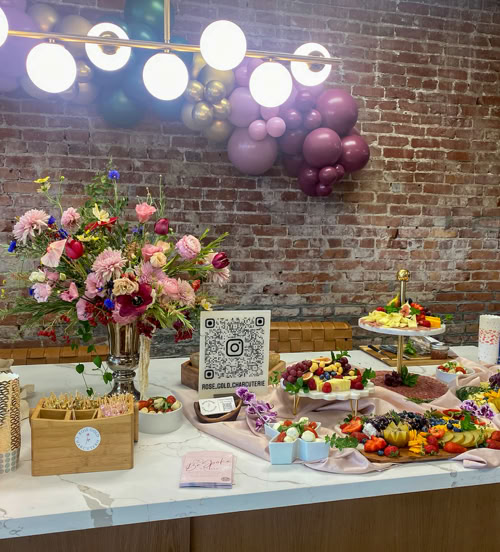 A beautifully styled charcuterie display on a marble counter featuring assorted grazing boards filled with fruits, cheeses, meats, and sweets. The setup includes fresh floral arrangements, a QR code for Rose Gold Charcuterie’s Instagram, a pink linen table runner, colorful balloons on a brick wall backdrop, and elegant lighting overhead.